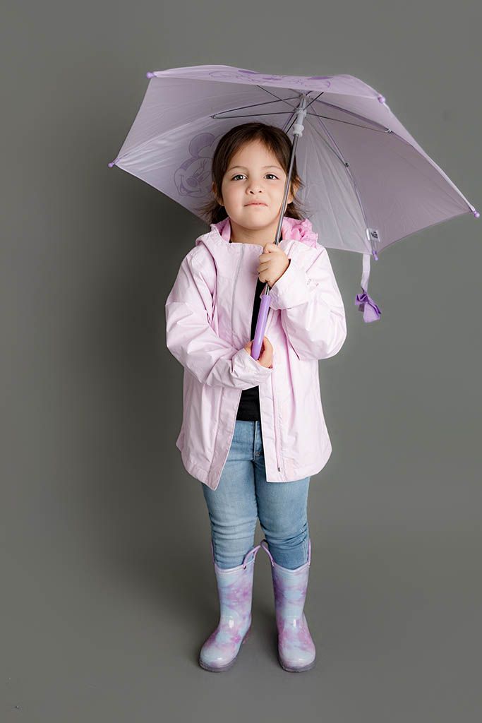 A young girl in a pink raincoat, jeans, and purple rain boots holds a lavender umbrella on a gray background.
