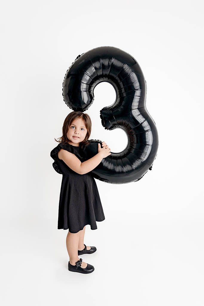 A little girl in a black dress holds a large black number 3 balloon, celebrating her third birthday on white background.
