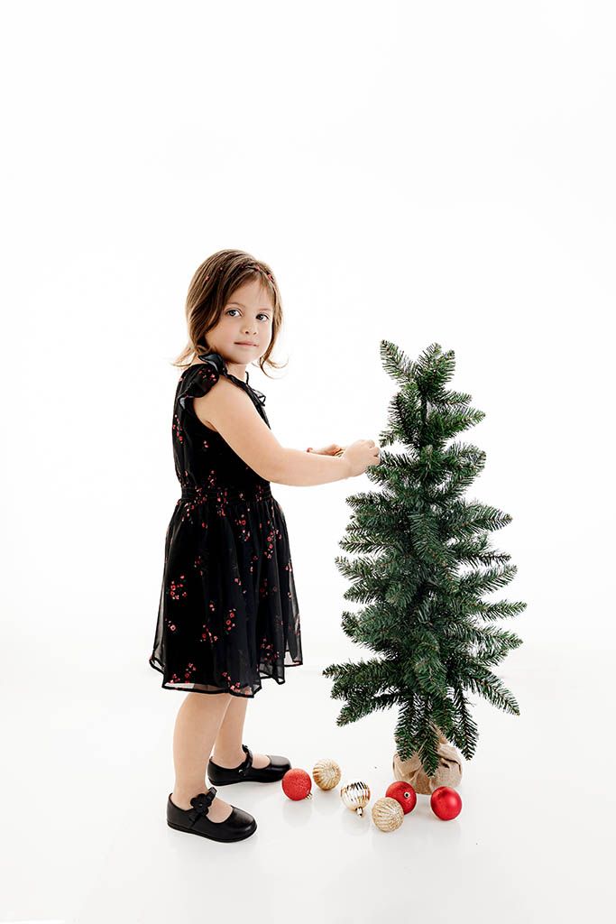 Young girl in black floral dress decorating a small Christmas tree with red and gold ornaments on white background.