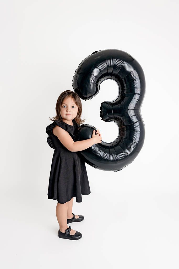A little girl in a black dress hugs a large black number 3 balloon against a white background.