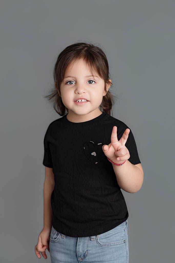 A cute toddler in a black t-shirt and jeans makes a peace sign gesture against a gray background.