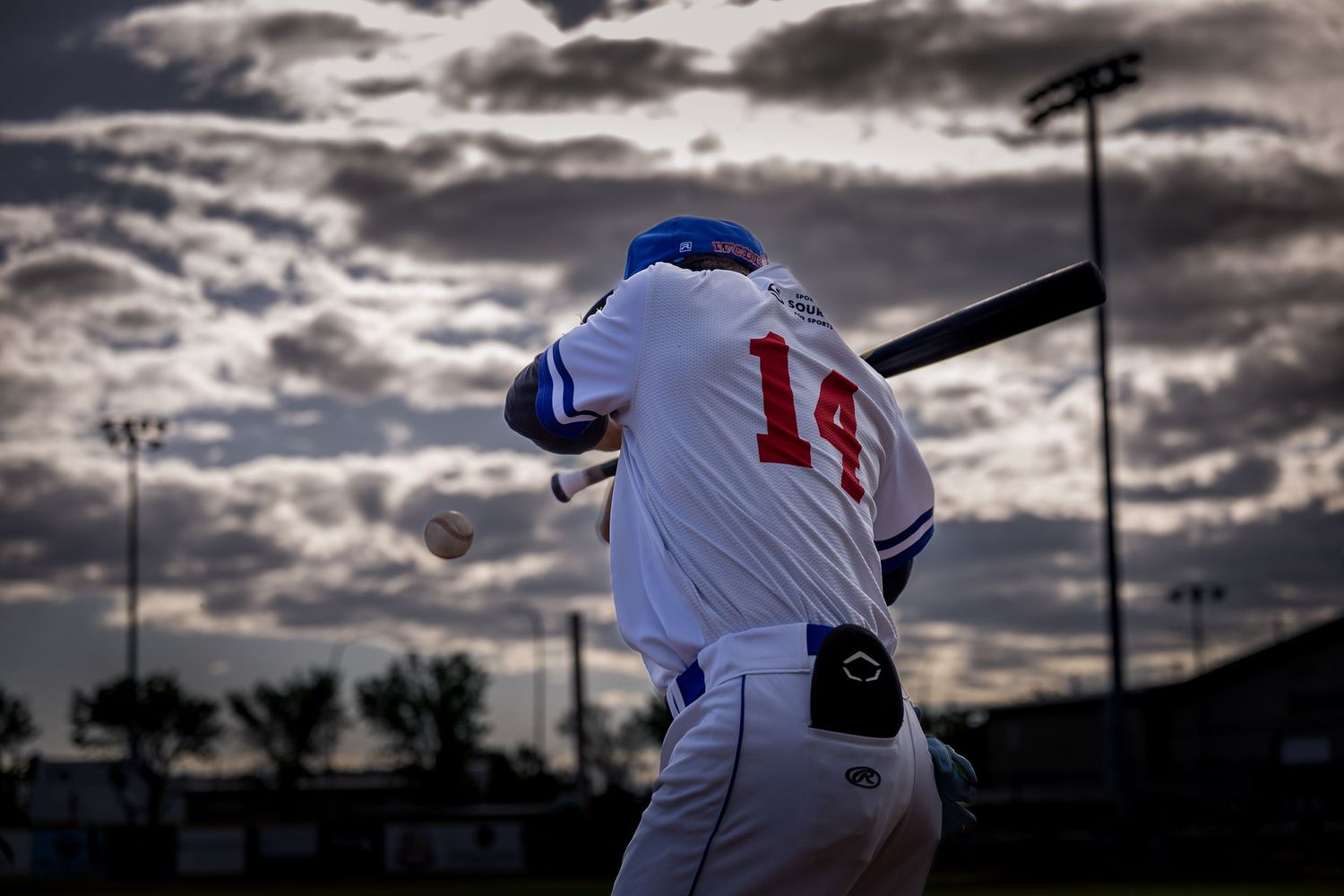 Brooks Bombers (WCBL) player number 14 swings bat under dramatic cloudy sky during evening game.
