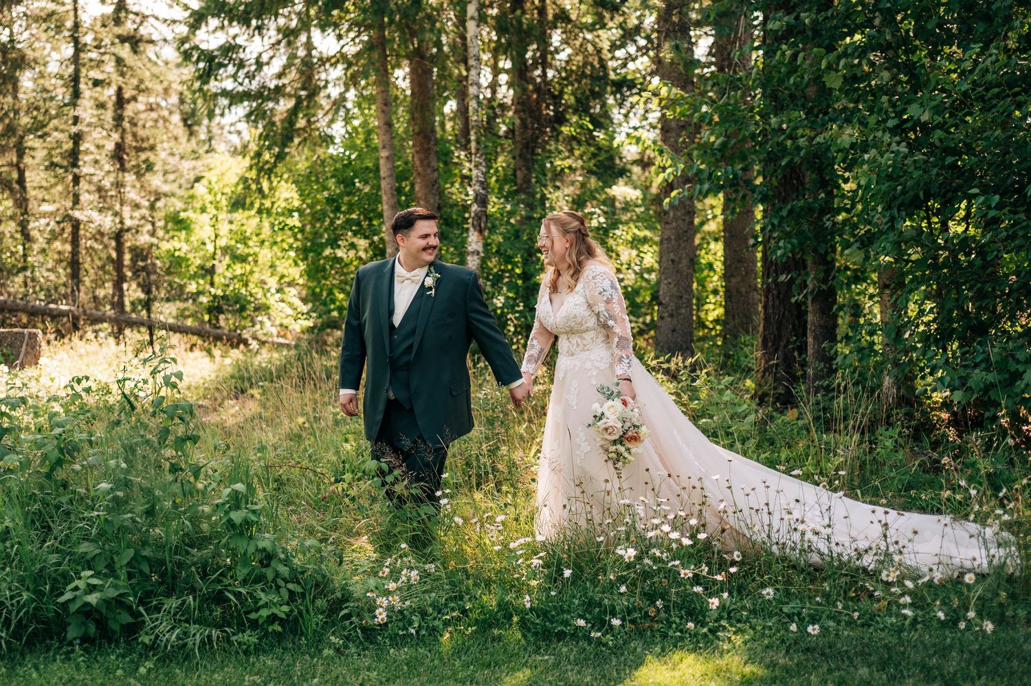 Bride and groom walk hand-in-hand through a lush green forest clearing, bride in lace gown with long train.
