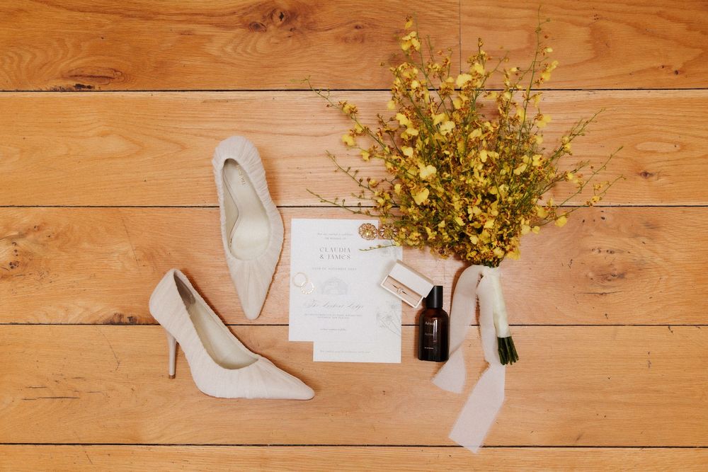 White bridal heels, wedding invitation, perfume bottles, and yellow wildflower bouquet arranged on rustic wooden floor.