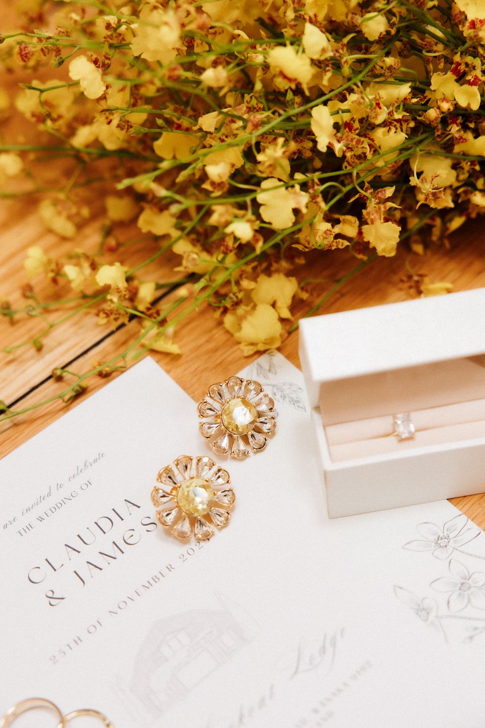 Gold floral earrings and diamond studs displayed on a wedding invitation beside yellow wildflowers on a wooden surface.