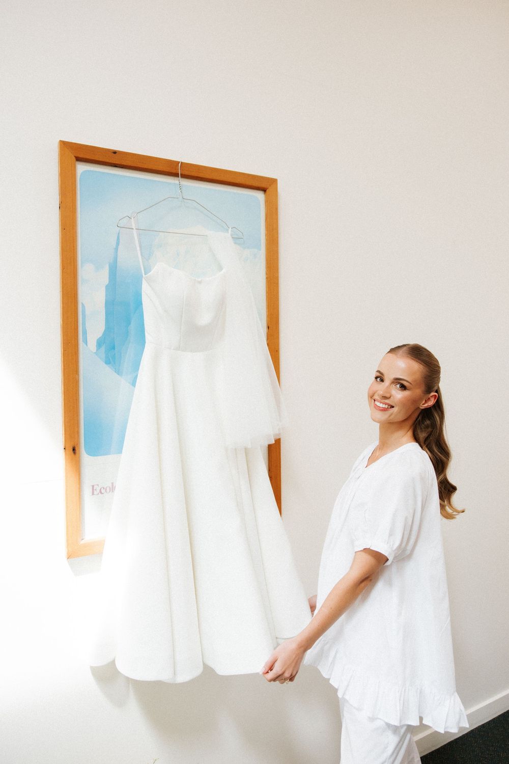 Smiling bride in white robe holding her elegant wedding dress hung on a frame in a bright room.