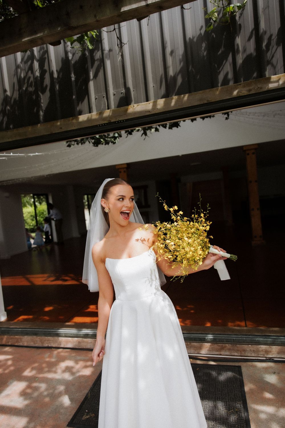 Excited bride in white strapless gown and veil holding yellow wildflower bouquet outside venue.