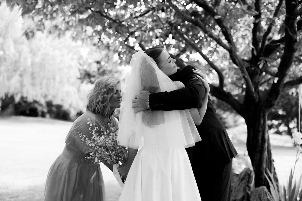 Bride shares emotional hug with guest under flowering tree at outdoor wedding.