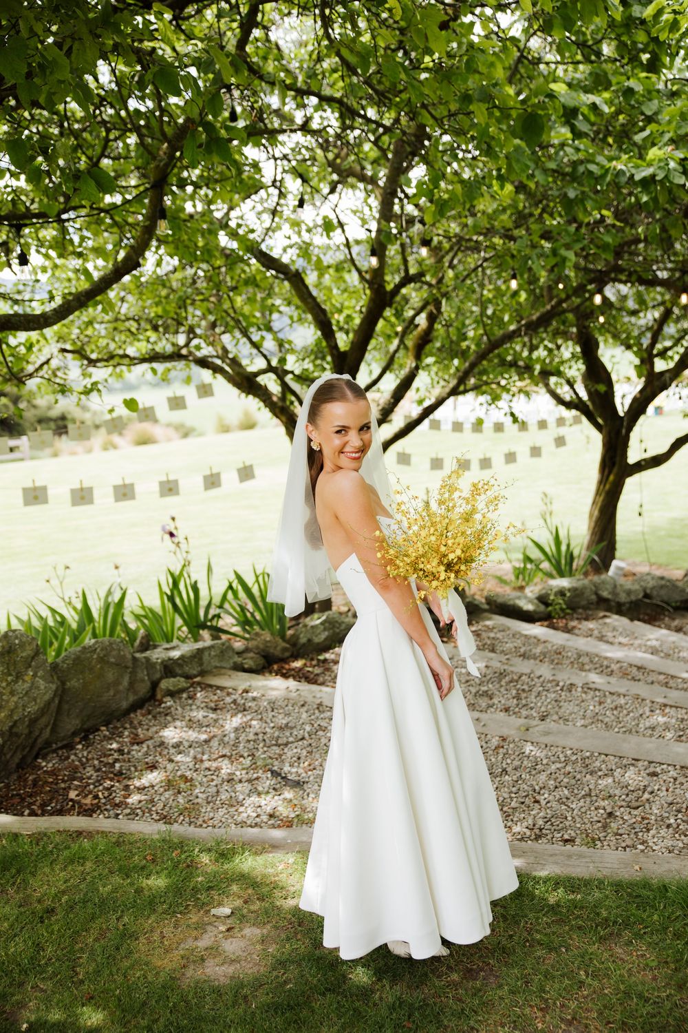 Smiling bride in white strapless gown holding yellow bouquet poses in lush garden setting. Wanaka Wedding