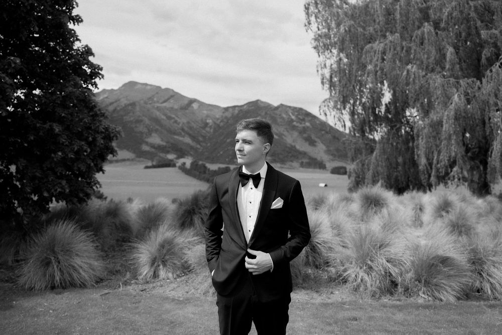 Dapper groom in tuxedo stands outdoors with mountain backdrop in black and white.