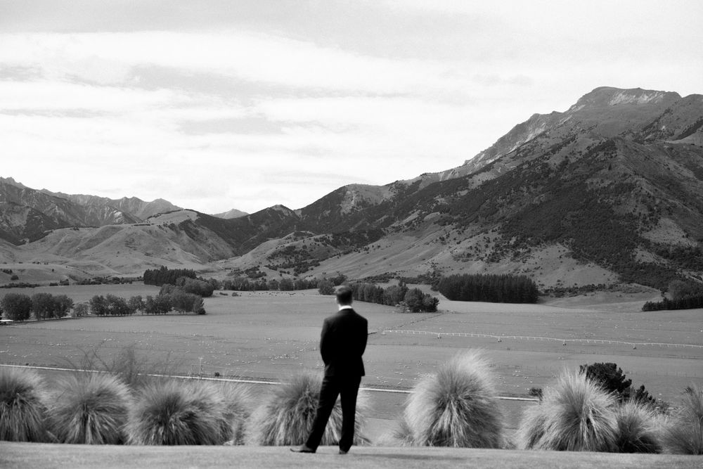 Groom stands alone gazing at stunning mountain valley landscape in black and white wedding photo.