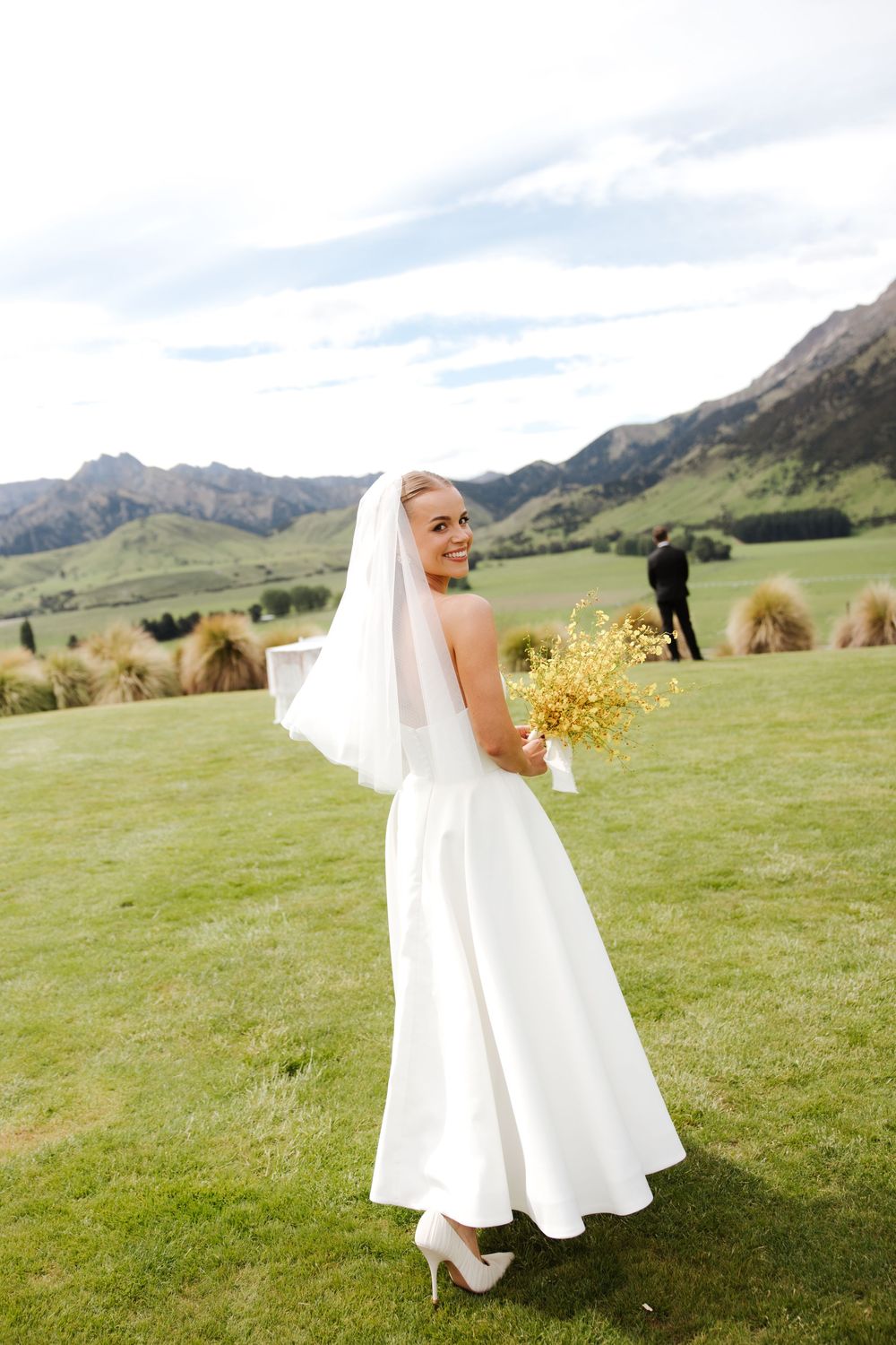 Smiling bride in white strapless gown and veil holding yellow bouquet with mountain landscape backdrop.