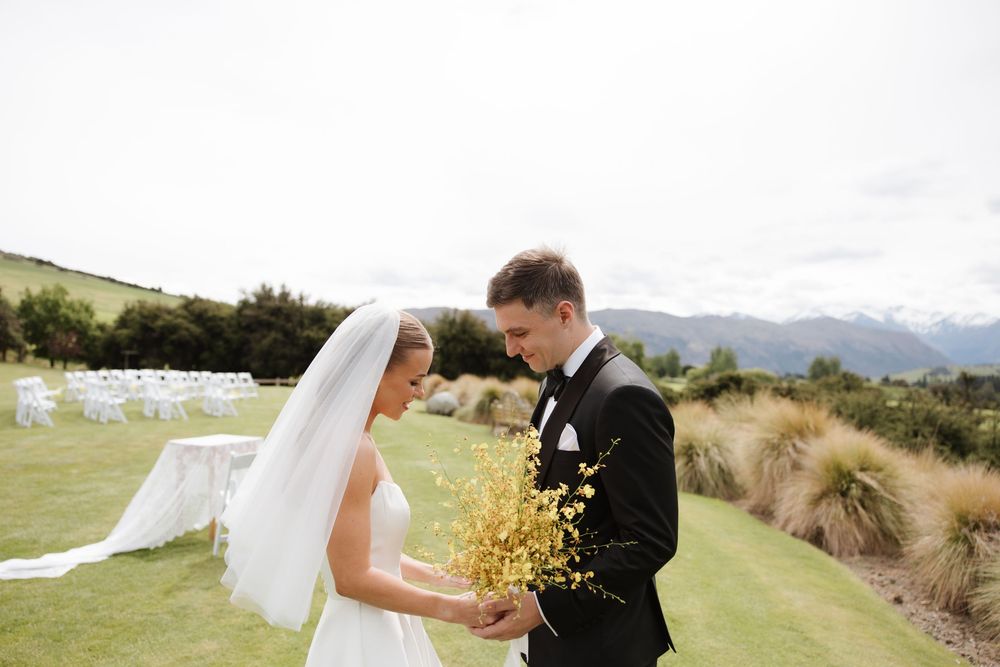 Bride and groom share a tender moment holding yellow wildflowers at an outdoor wedding venue with mountain views.