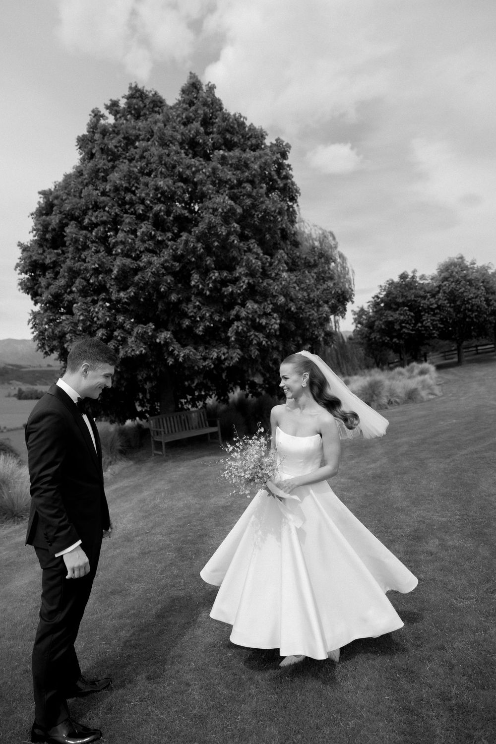 Black and white photo of bride and groom sharing a joyful moment on a green lawn with a large tree backdrop.