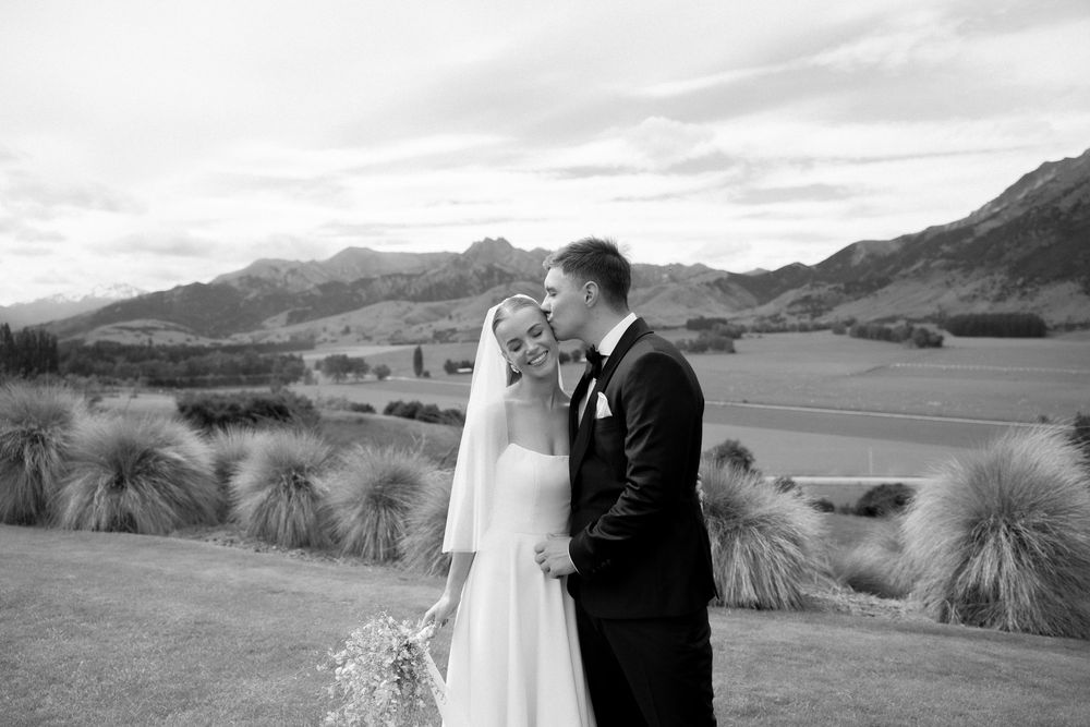Bride and groom embrace smiling before scenic mountain backdrop in elegant black and white portrait. Wanaka