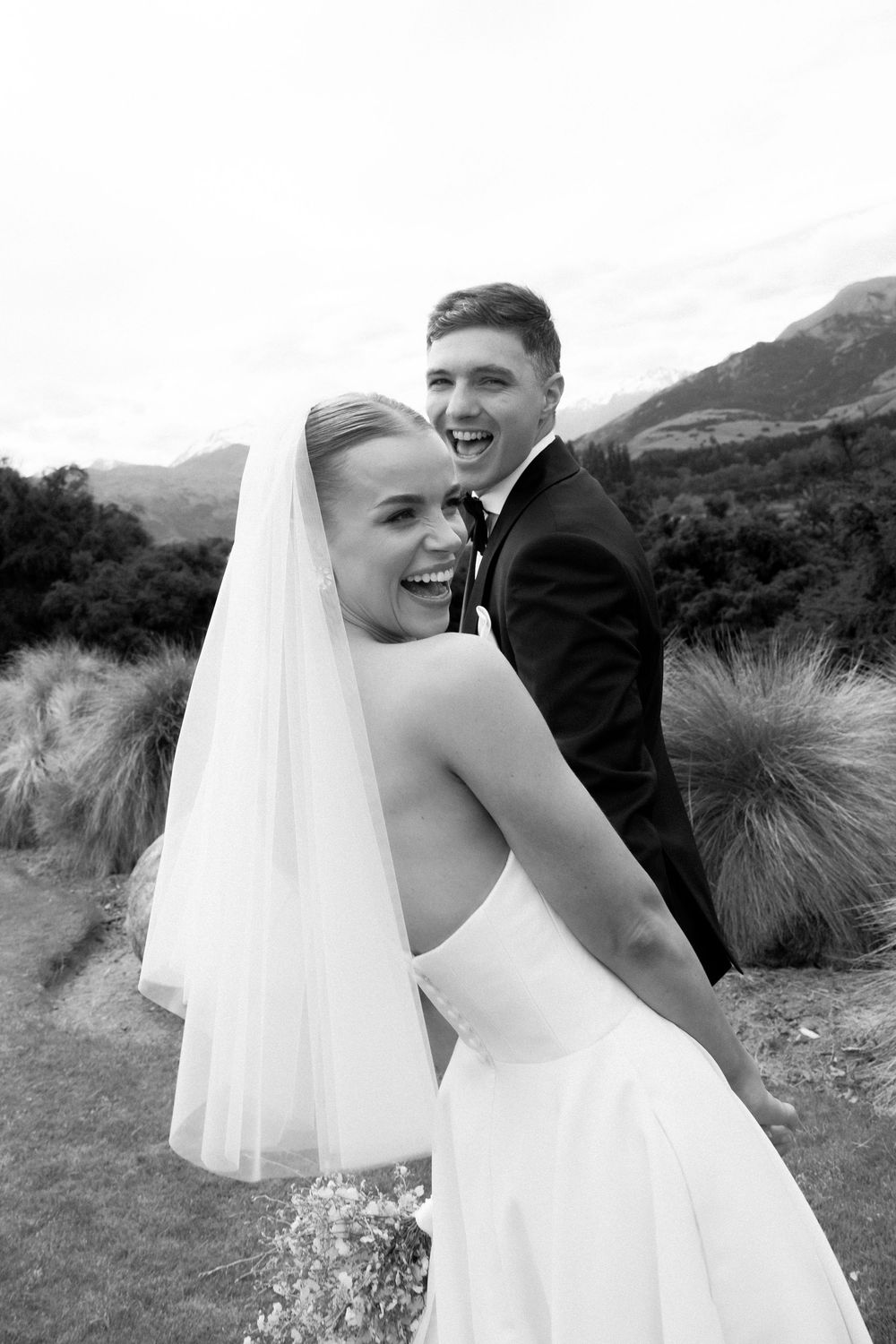 Joyful bride and groom laughing together in black and white portrait with mountain scenery behind them.
