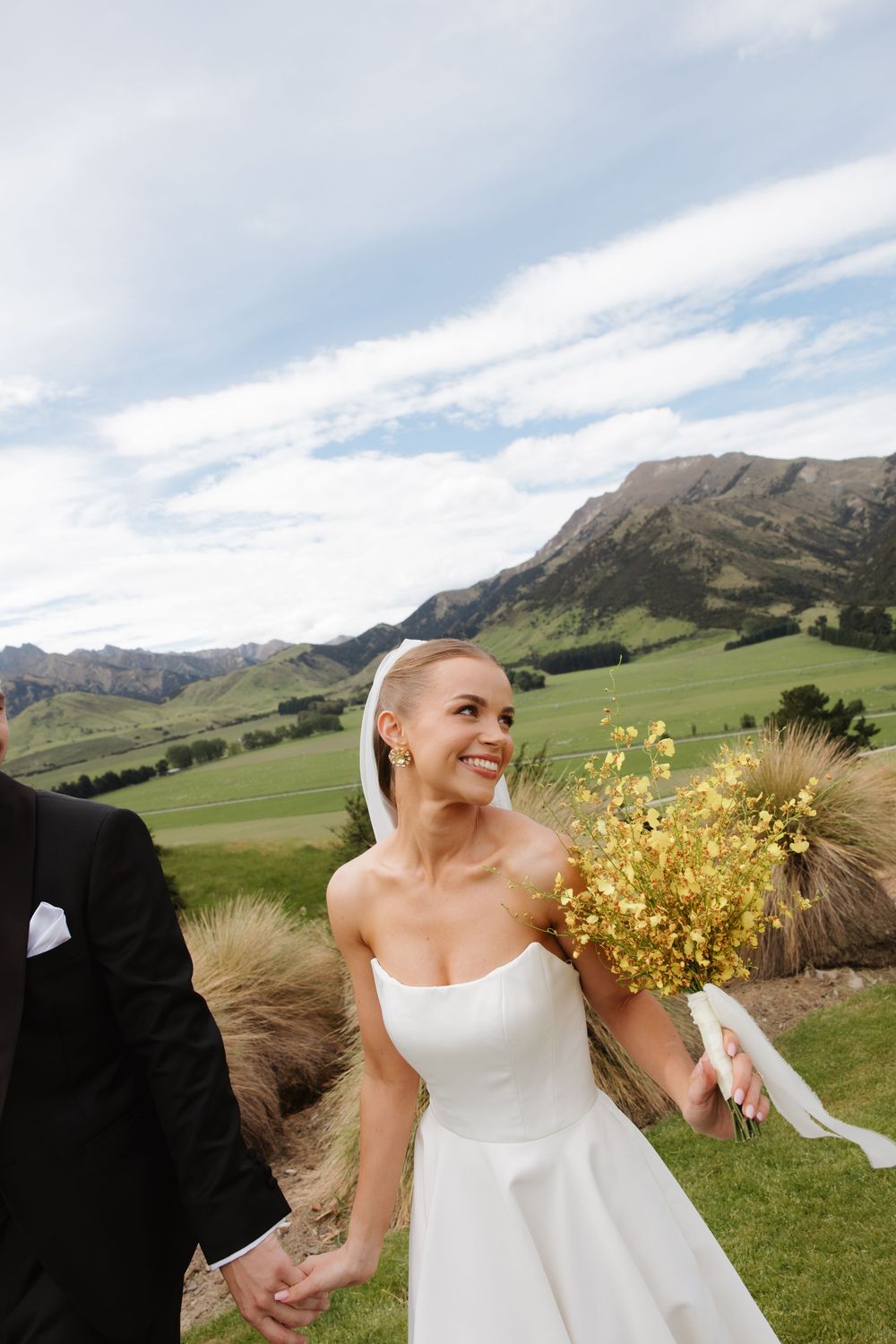 Smiling bride in strapless white gown holding yellow bouquet with groom against scenic mountain landscape.