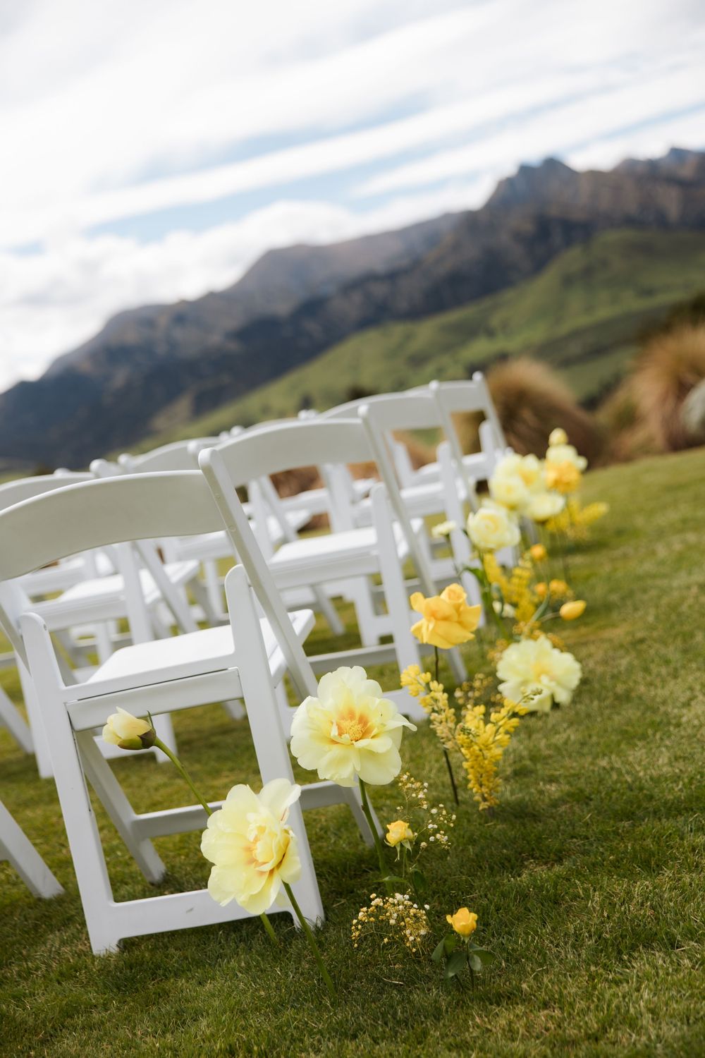 White folding chairs line a grass aisle decorated with yellow flowers at an outdoor wedding ceremony with mountain views.