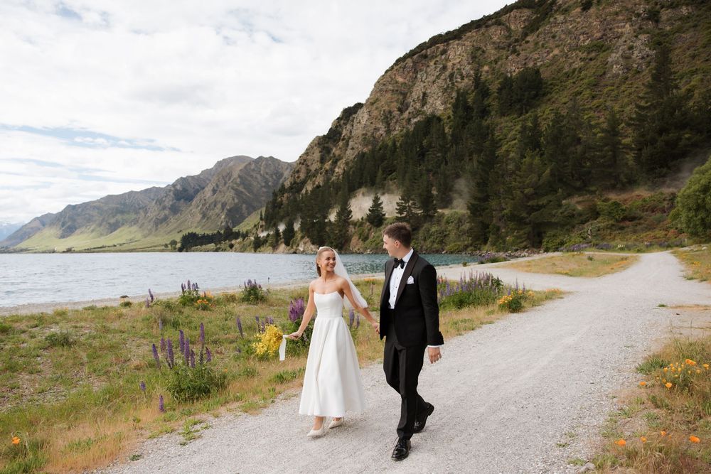 Bride and groom walking hand in hand along a scenic lakeside gravel path with mountain backdrop in New Zealand.