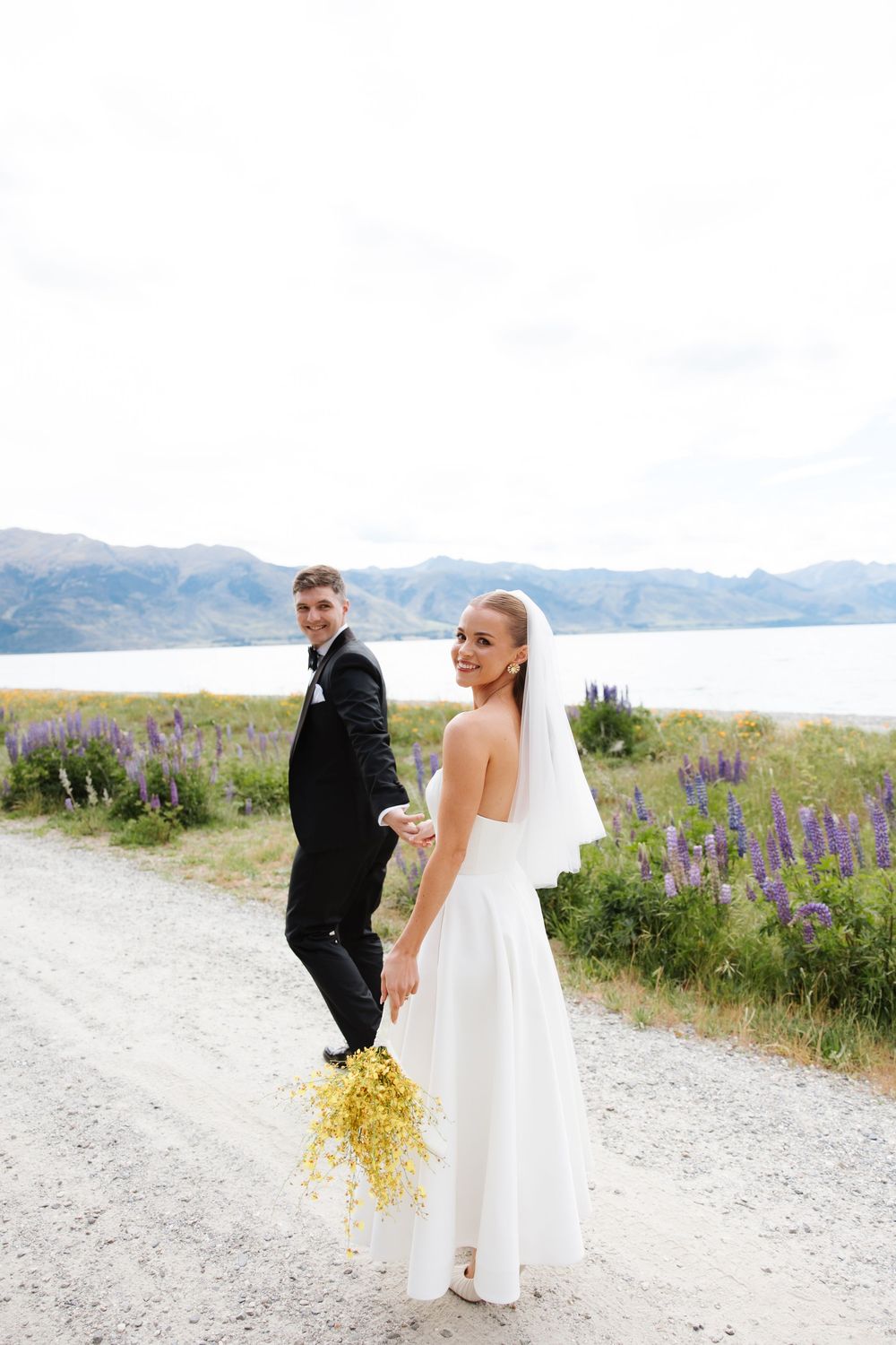 Bride and groom holding hands on a scenic gravel path surrounded by lupins with a mountain lake backdrop.