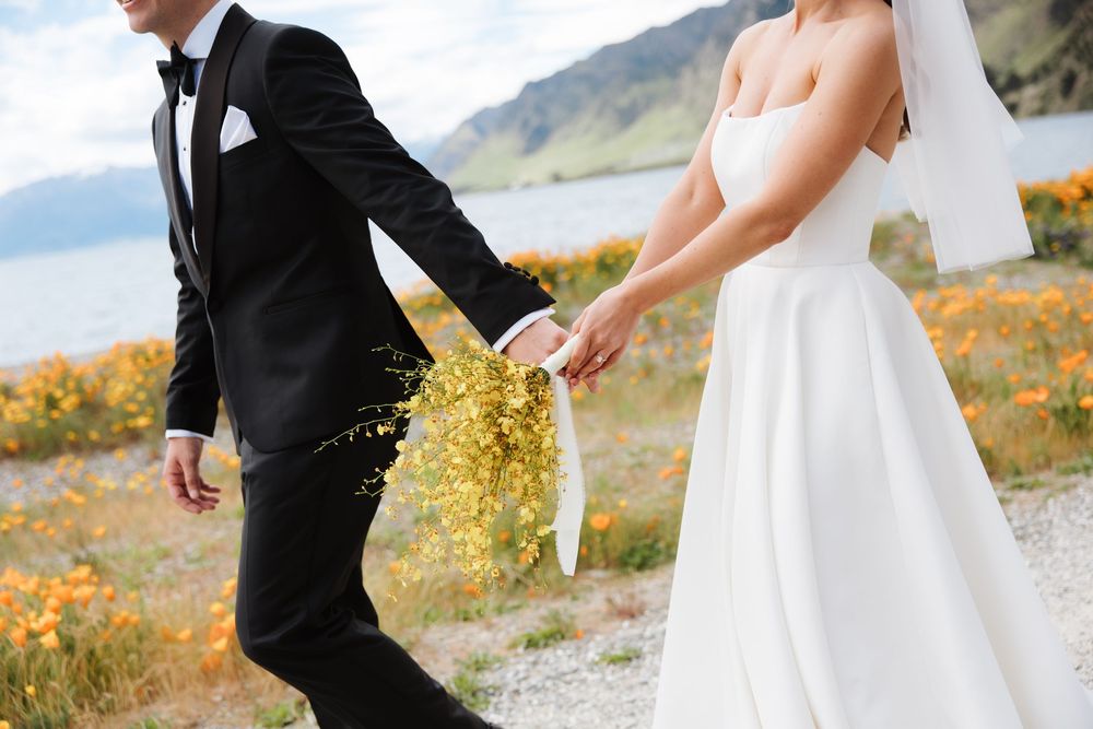 Wedding couple walking hand in hand near lake with orange wildflowers, bride holding yellow bouquet.