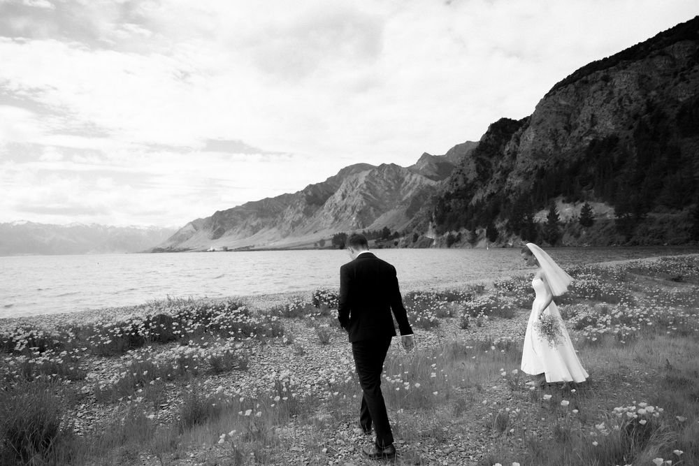 Bride and groom walk through wildflower field toward dramatic lakeside cliffs.