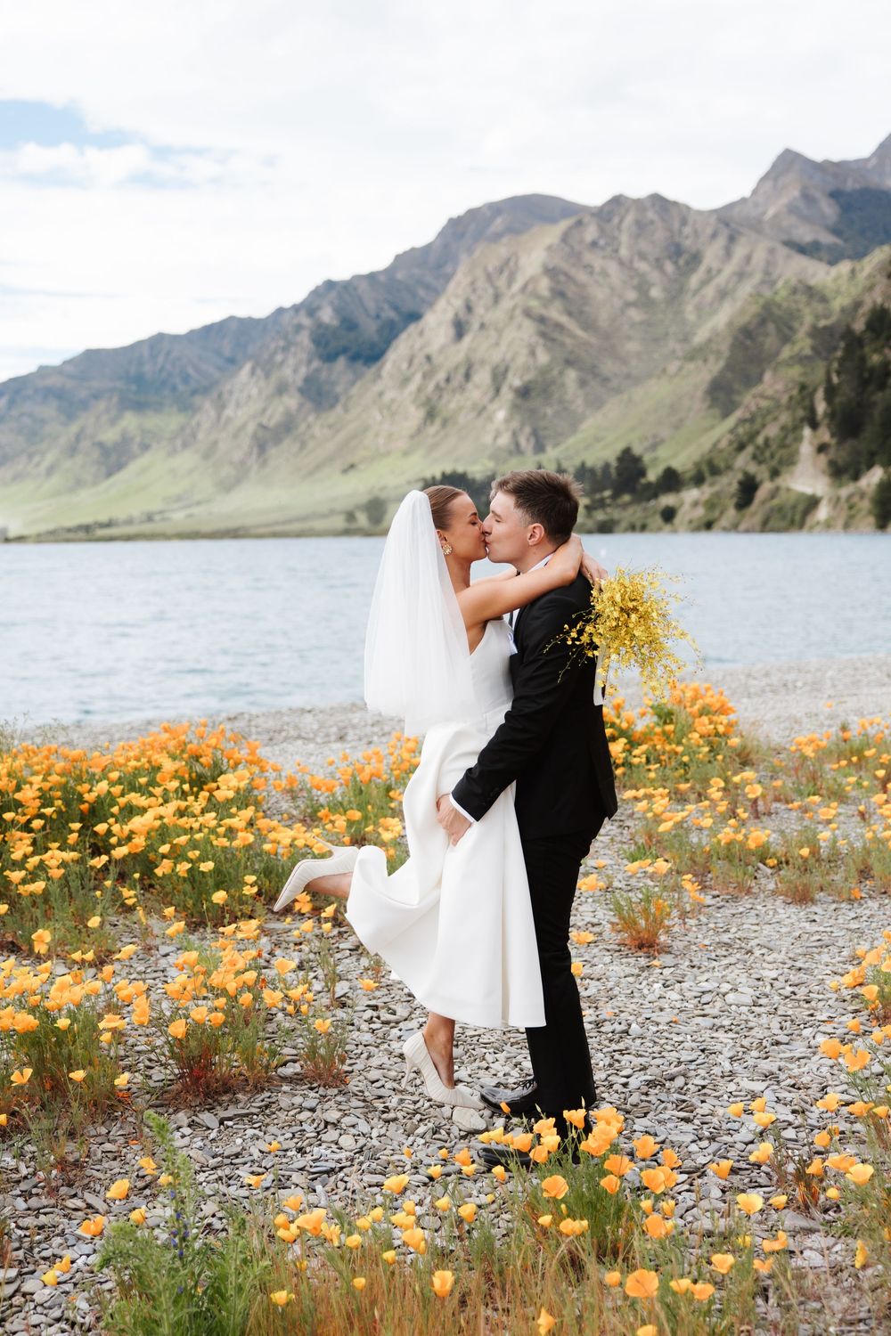Groom lifting bride for a kiss among orange wildflowers with a stunning mountain lake in the background. Lake Hāwea