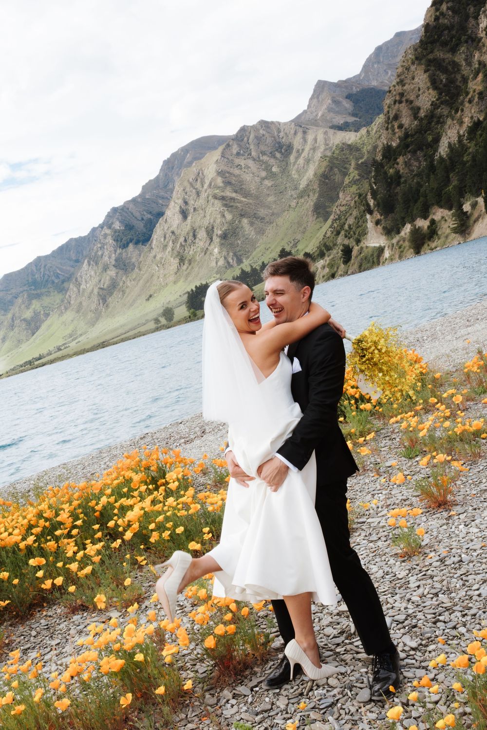 Joyful bride and groom embracing by a mountain lake surrounded by vibrant orange wildflowers.