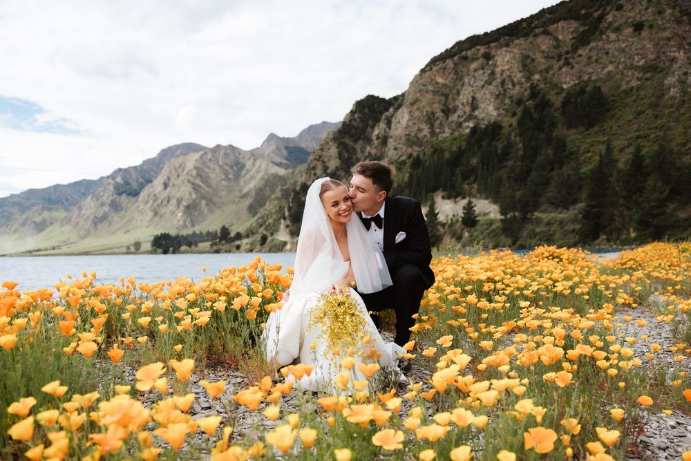 Wedding couple poses romantically among vibrant yellow wildflowers with mountain lake scenery behind them.