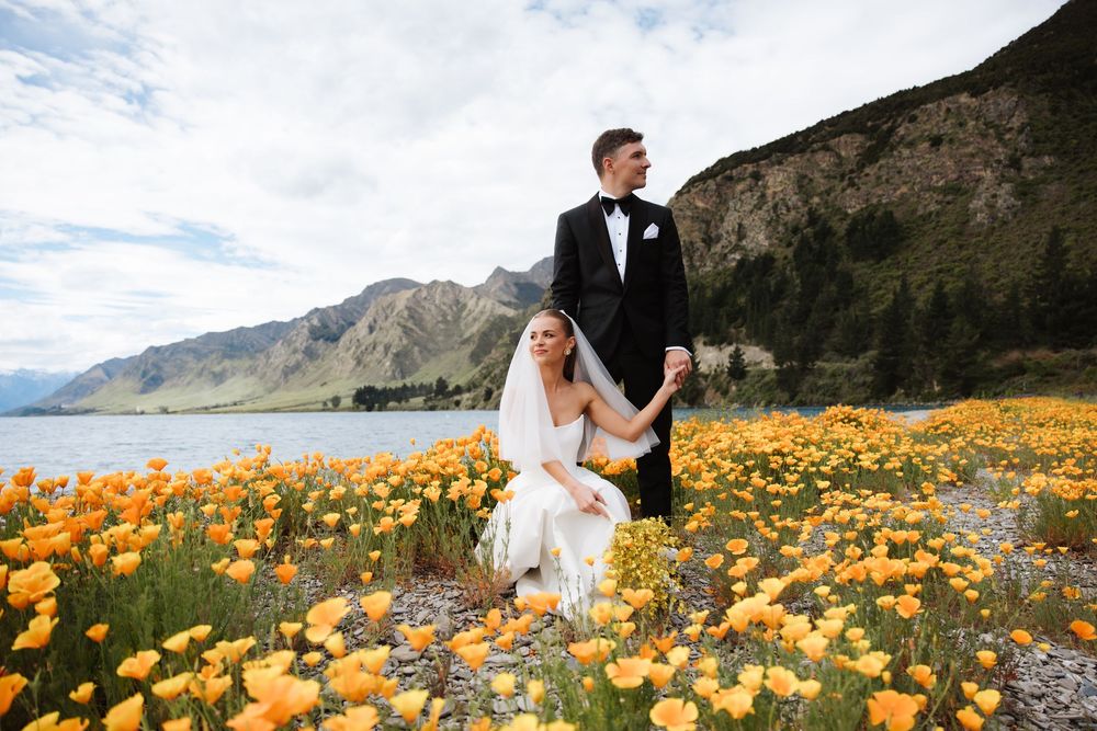 Wedding couple posing in field of orange poppies with mountain lake backdrop in New Zealand. Wanaka Wedding photography