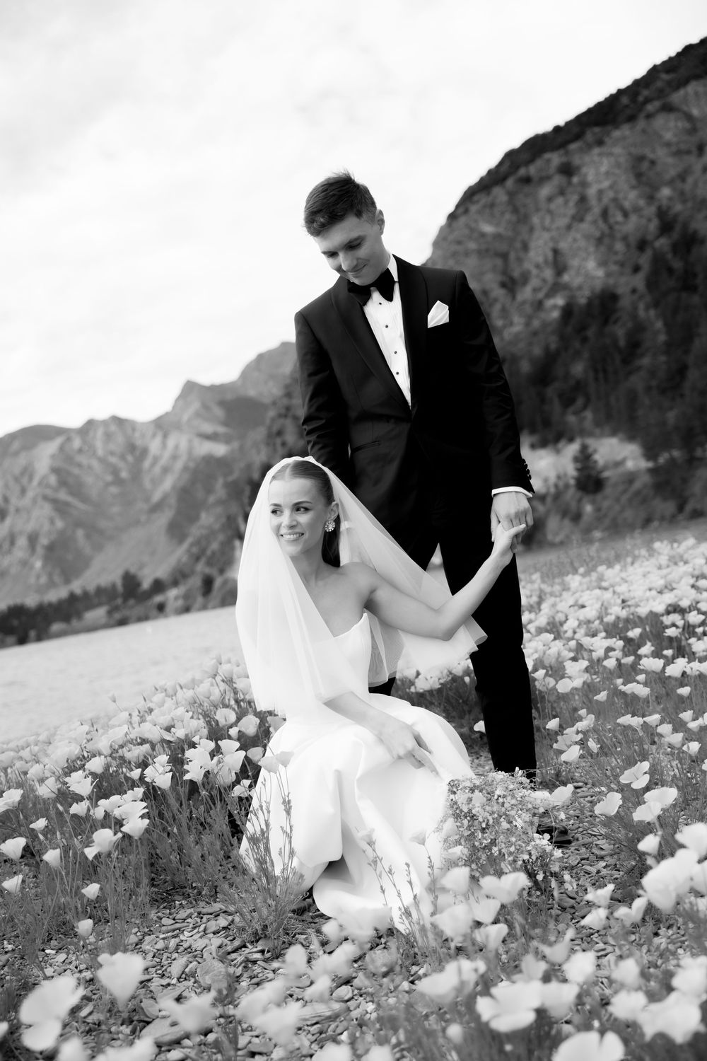 Black and white photo of bride sitting in wildflower field holding grooms hand with mountains in background.