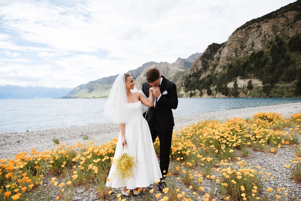 Groom kissing bride's hand in orange poppy field by lake with mountain scenery behind them.