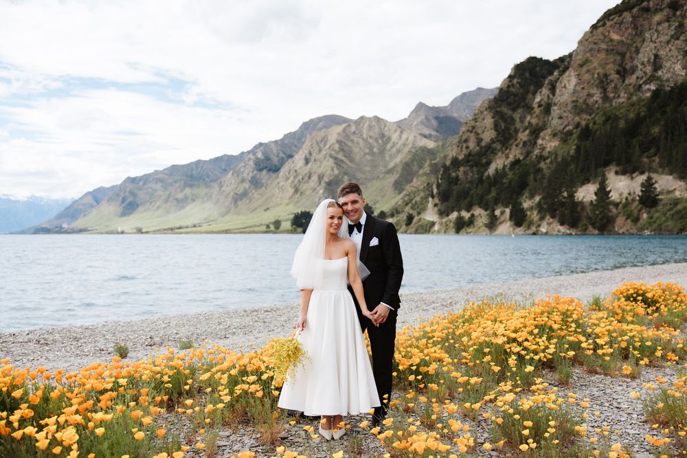 Bride and groom posing together by a scenic mountain lake with bright orange poppies in bloom.