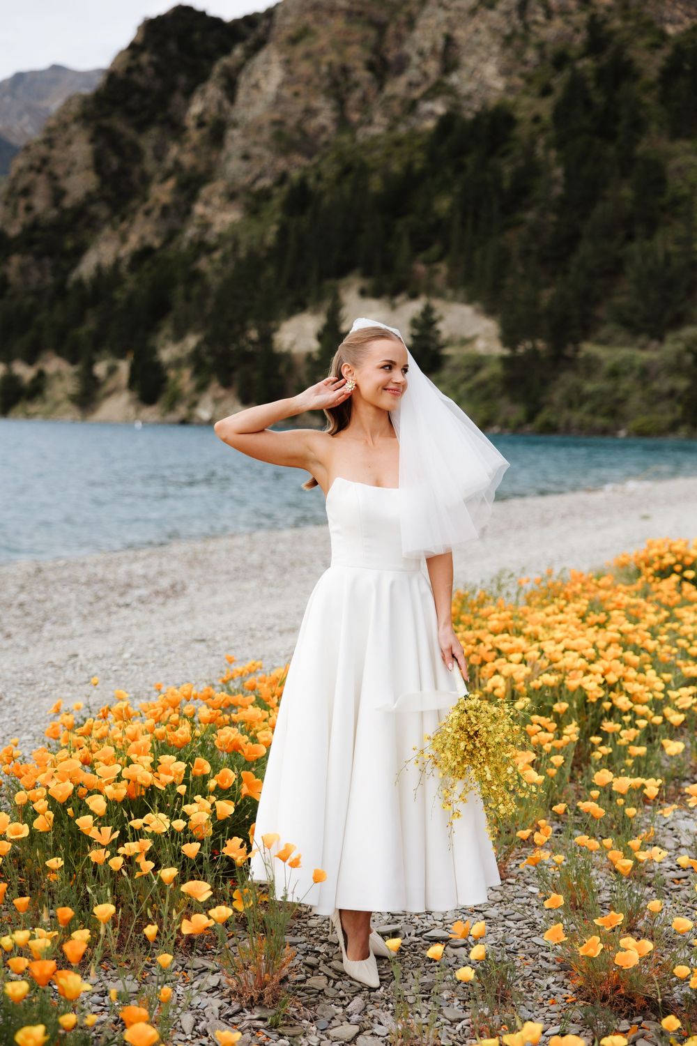 Smiling bride in white tea-length gown and veil standing among orange wildflowers by a mountain lake.