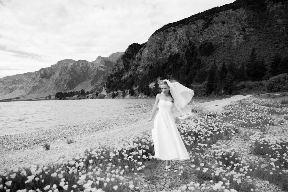Bride in white gown and veil stands in wildflower field with mountain backdrop in black and white.