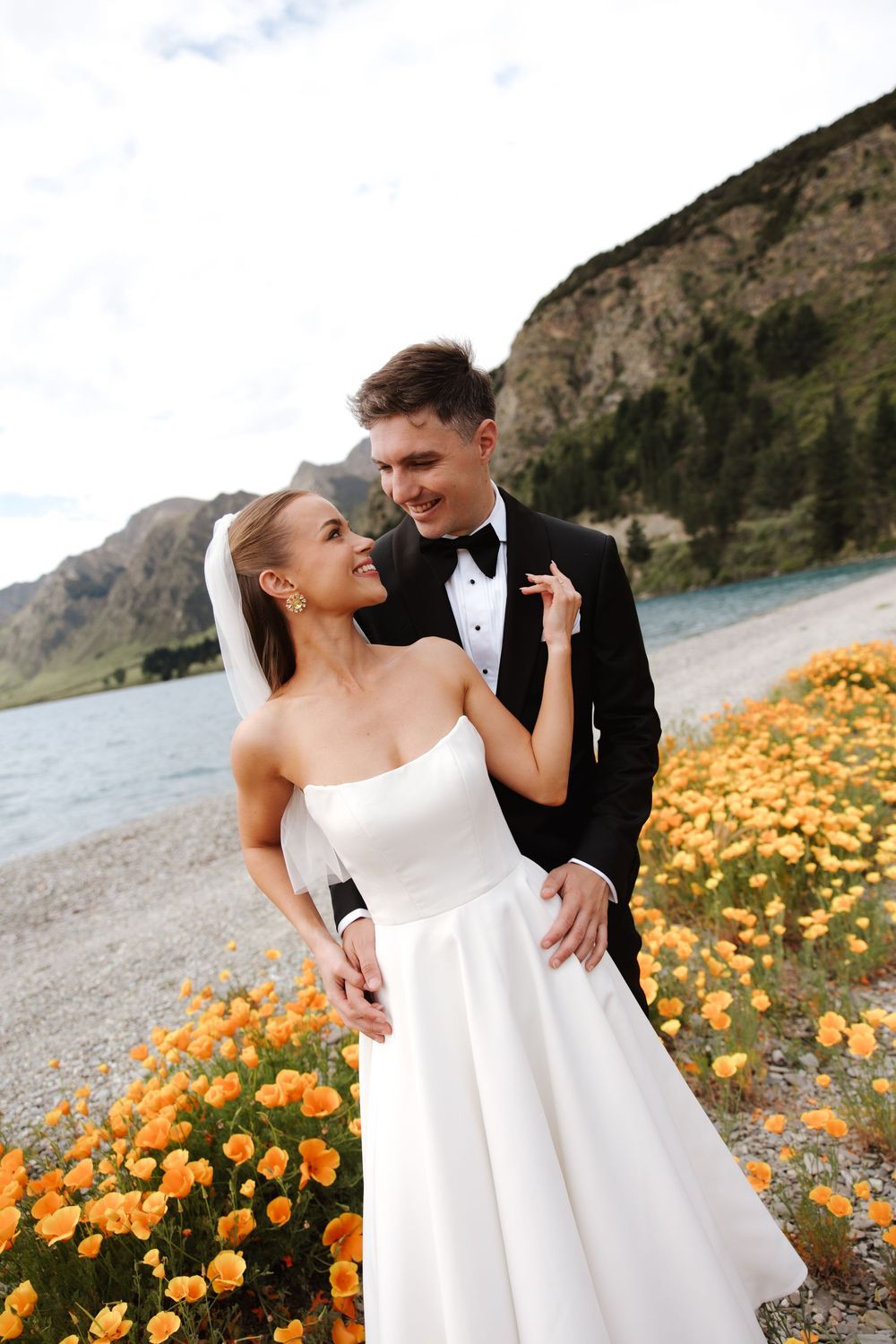 Bride and groom embracing among orange poppies with stunning mountain lake backdrop on wedding day.