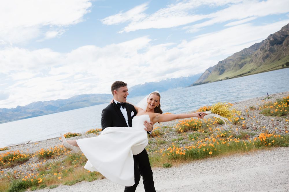 Groom carries laughing bride along scenic lakeside with mountain views.