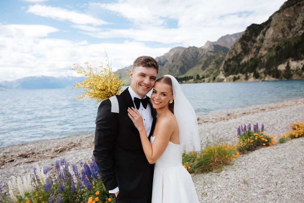 Newlywed couple embracing on lakeside shore with mountains and wildflowers in background.