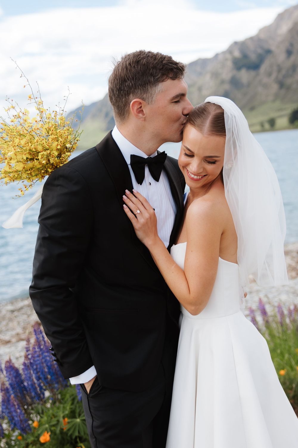Groom kissing bride on the forehead near a lakeside garden with purple lupins and mountains behind them.