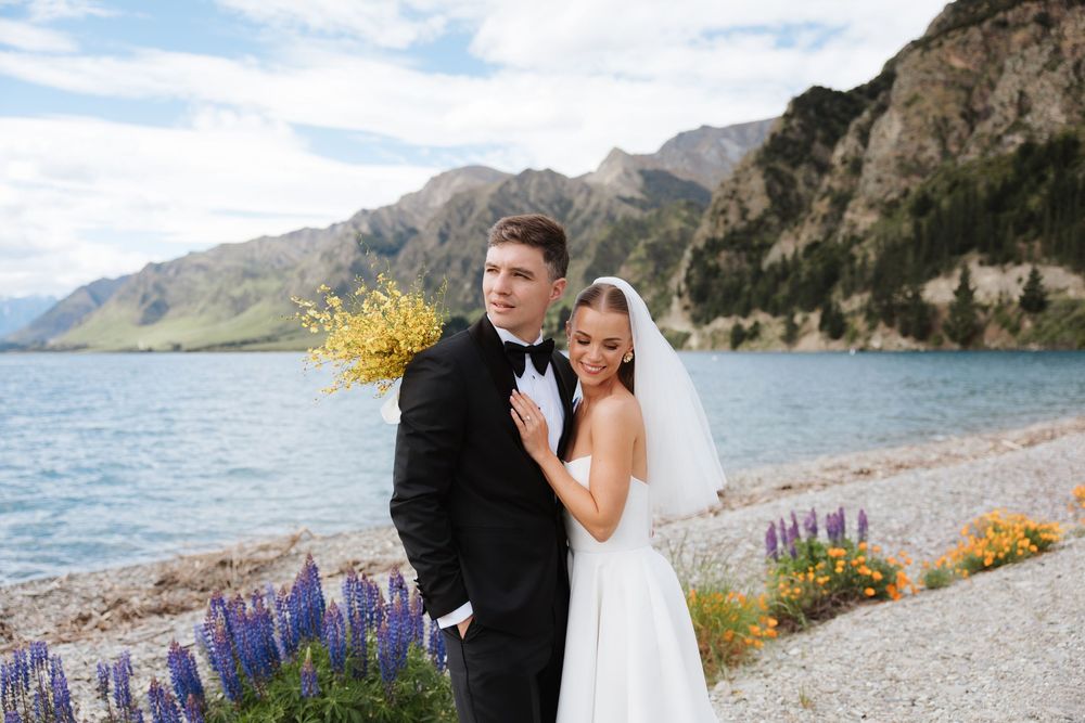Bride and groom posing by a mountain lake with lupine flowers in Wanaka, New Zealand.