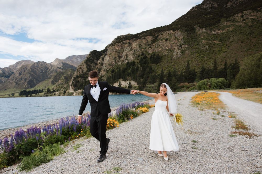 Newlyweds strolling along lakeside gravel path with purple lupins and mountains in New Zealand.