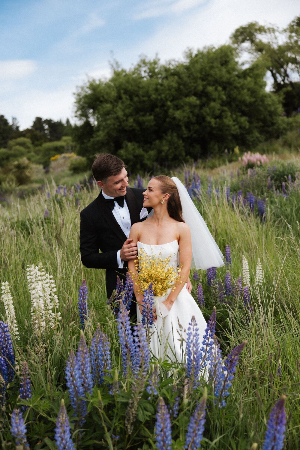 Bride and groom embrace among purple lupine wildflowers, bride holding yellow bouquet, groom in black tuxedo.