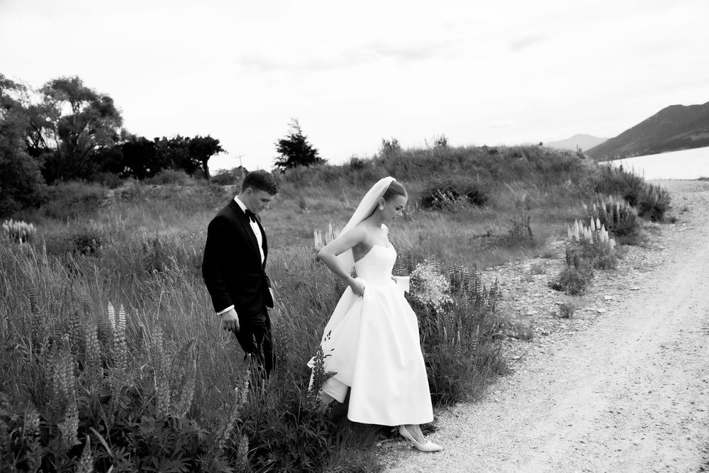 Black and white photo of bride and groom walking along a rural path surrounded by tall grasses.