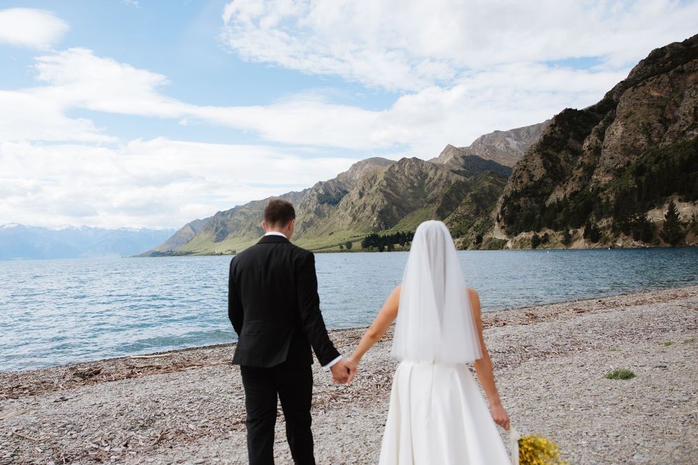 Bride and groom holding hands facing a stunning turquoise mountain lake on a rocky shoreline.