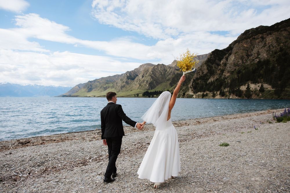 Bride and groom holding hands on rocky lakeside beach with mountain backdrop, bride raising yellow bouquet.
