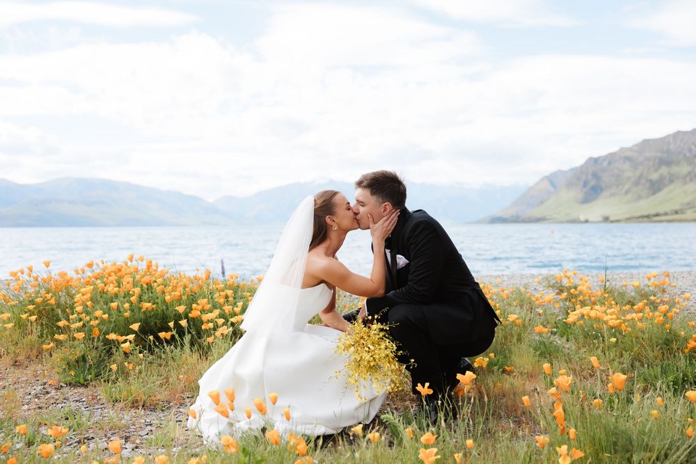 Bride and groom sharing a romantic kiss among orange wildflowers with a Lake Hāwea mountain lake backdrop.