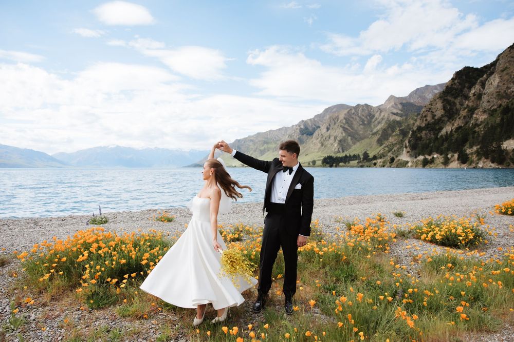 Groom twirling bride in white dress among orange poppies on rocky lakeshore with mountains.