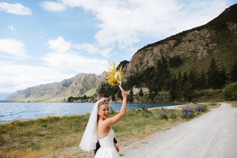 Joyful bride tossing yellow bouquet in the air on a gravel path beside a scenic mountain lake.