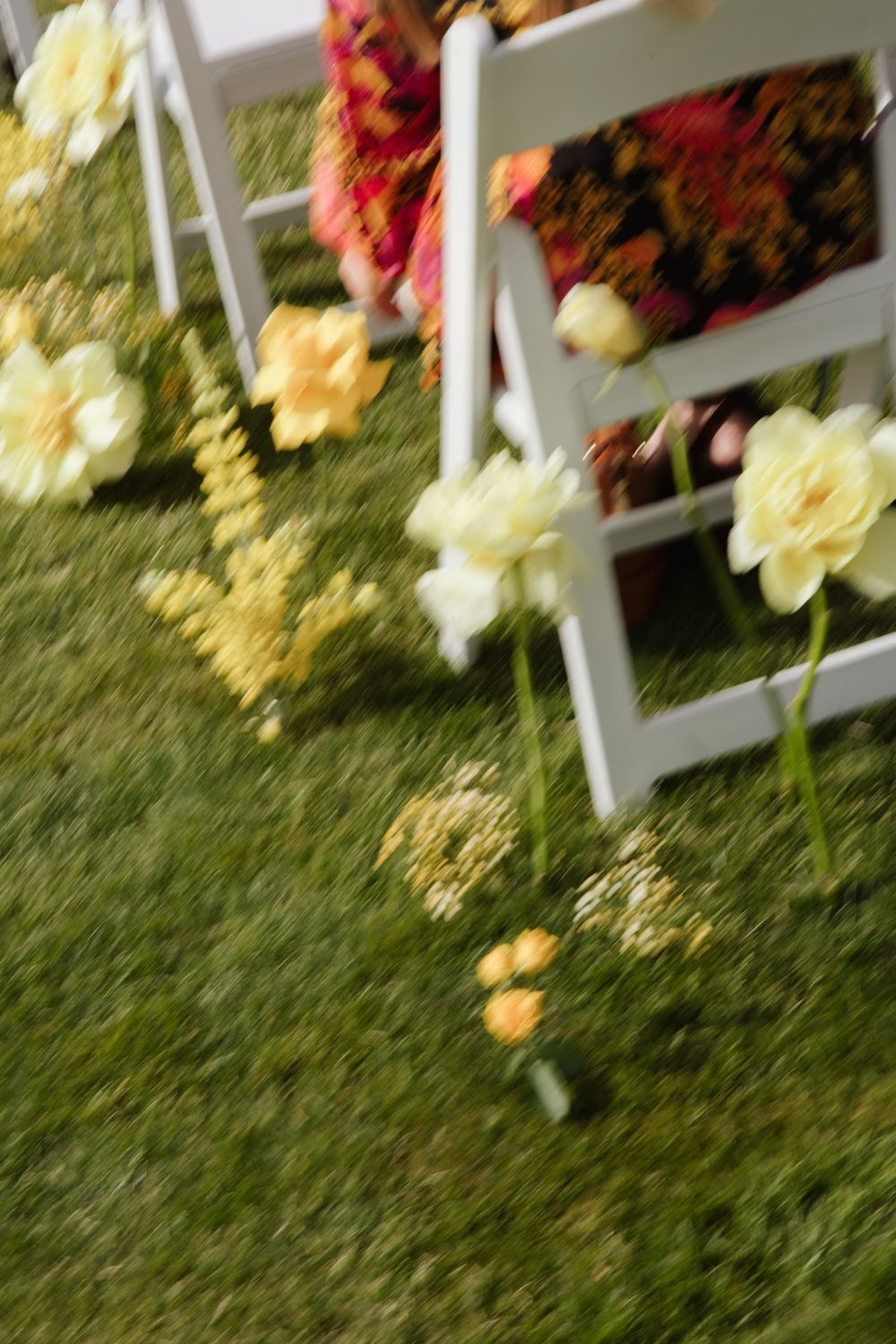 Blurred artistic shot of yellow and white flowers scattered on grass beside white wedding chairs.