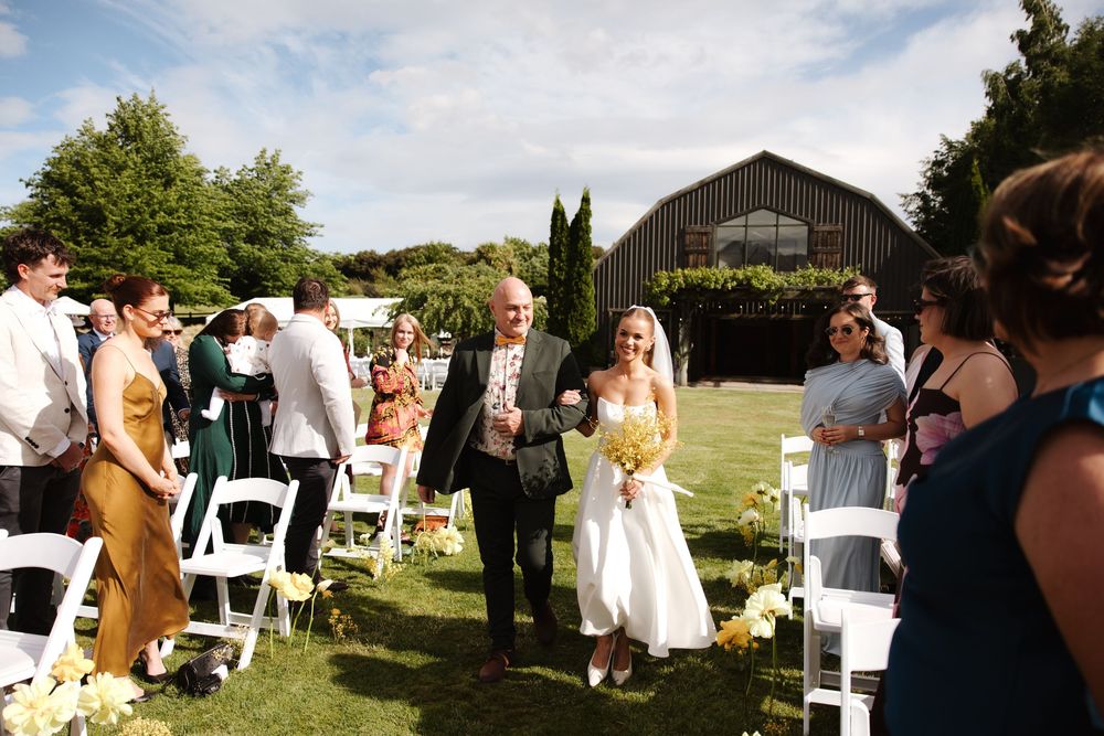 Bride walks down the aisle with escort at an outdoor wedding ceremony beside a rustic barn venue.